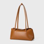A cognac brown vegan leather baguette shoulder bag with long slim straps and a softly curved top, shown on a white background. Additional product view 4.