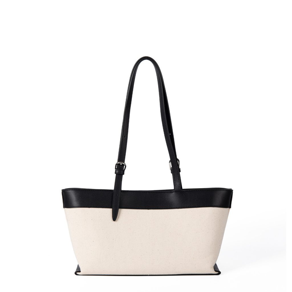 An off-white canvas tote with black vegan leather top trim, base, and adjustable shoulder straps.