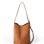 A tall cognac brown faux suede bucket bag with an adjustable vegan leather shoulder strap hanging against a white backdrop.