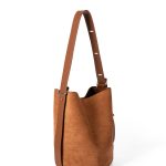 A tall cognac brown faux suede bucket bag with an adjustable vegan leather shoulder strap hanging against a white backdrop. Additional product view 3.