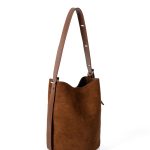 A tall cognac brown faux suede bucket bag with an adjustable vegan leather shoulder strap hanging against a white backdrop. Additional product view 4.