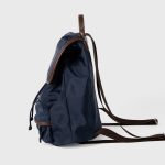 A black nylon backpack with cognac brown vegan leather piping, a top flap over a drawstring, and two lower front pockets. Additional product view 4.