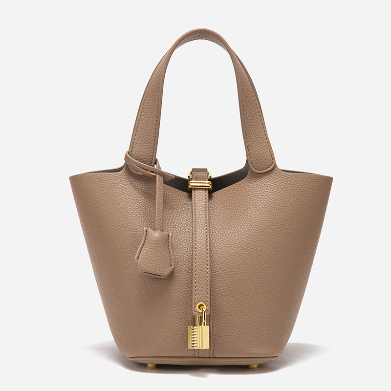 A taupe pebbled vegan leather bucket bag with dual top handles, a gold padlock on the center strap, and a matching clochette.
