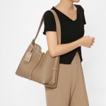 A taupe pebble-grain vegan leather hobo worn on the shoulder, showing gold hardware and a striped interior lining.